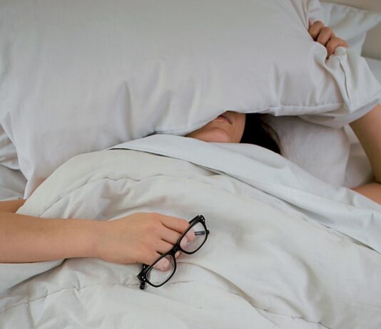 피로를 극복하는 에너지 보충법 person lying on bed while covering face with pillow and holding eyeglasses