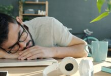 ‘스트레스 리커버리’ 시대 — 번아웃을 예방하는 5분 루틴 Man sleeping at desk with headphones and coffee.