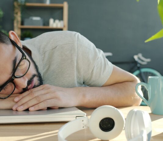 ‘스트레스 리커버리’ 시대 — 번아웃을 예방하는 5분 루틴 Man sleeping at desk with headphones and coffee.
